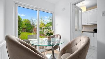 A modern living room with a glass table, two beige armchairs, and a view of a garden through large sliding doors.