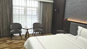 A hotel room with a bed, two armchairs, a small table, a wall clock, and a window with curtains.