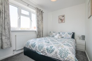 A bedroom with a bed, a radiator, a window with curtains, a bedside table, and a wall-mounted picture.