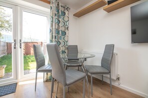 A dining area with a glass table and four grey chairs, a TV mounted on the wall, and a view of the outside through a glass door.