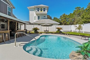 A swimming pool with a curved edge, surrounded by a deck area with a white fence, a covered seating area, and a building with a two-story section and a balcony. - Stylish Panama City Home w/ Pool, Tiki Bar, & Walkable Beach Access (Panama City Beach)