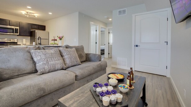A living room with a sofa, a coffee table with snacks and drinks, a kitchen in the background, and a TV mounted on the wall. - The Gameday Landing at Stadium Place (Manhattan)
