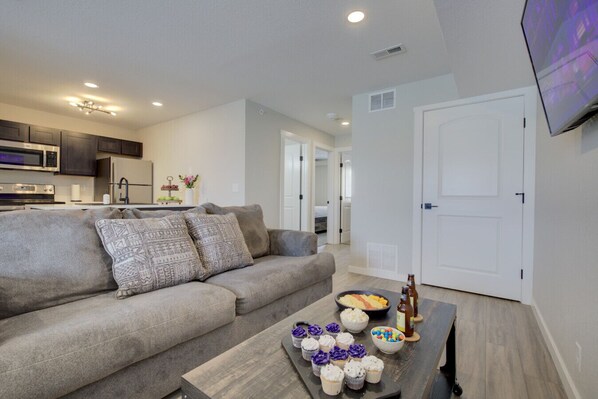 A living room with a sofa, a coffee table with snacks and drinks, a kitchen in the background, and a TV mounted on the wall.