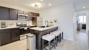 A modern kitchen with dark cabinets, stainless steel appliances, a central island with bar stools, and a view into the adjacent dining area. - The Gameday Landing at Stadium Place (Manhattan)