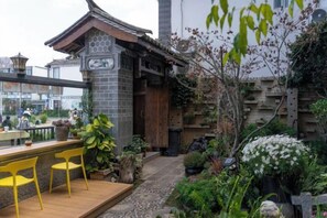 Een traditioneel Japans gebouw met een houten terras, gele stoelen en een tuin met diverse planten en een stenen lantaarn.