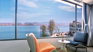 A modern hotel room with a large window offering a scenic view of a lake, a pier, and distant mountains. There are two chairs and a small table with a vase of flowers. - Longkan Yunding Courtyard Hotel (Dali City)