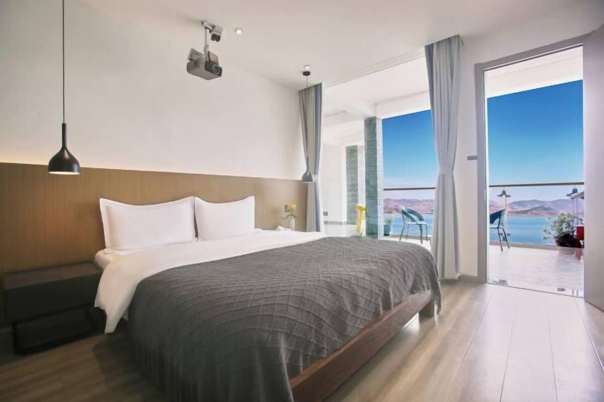Quarto de hotel moderno com uma cama grande, uma mesa de cabeceira, uma luminária pendente e vista para o mar através de uma varanda.