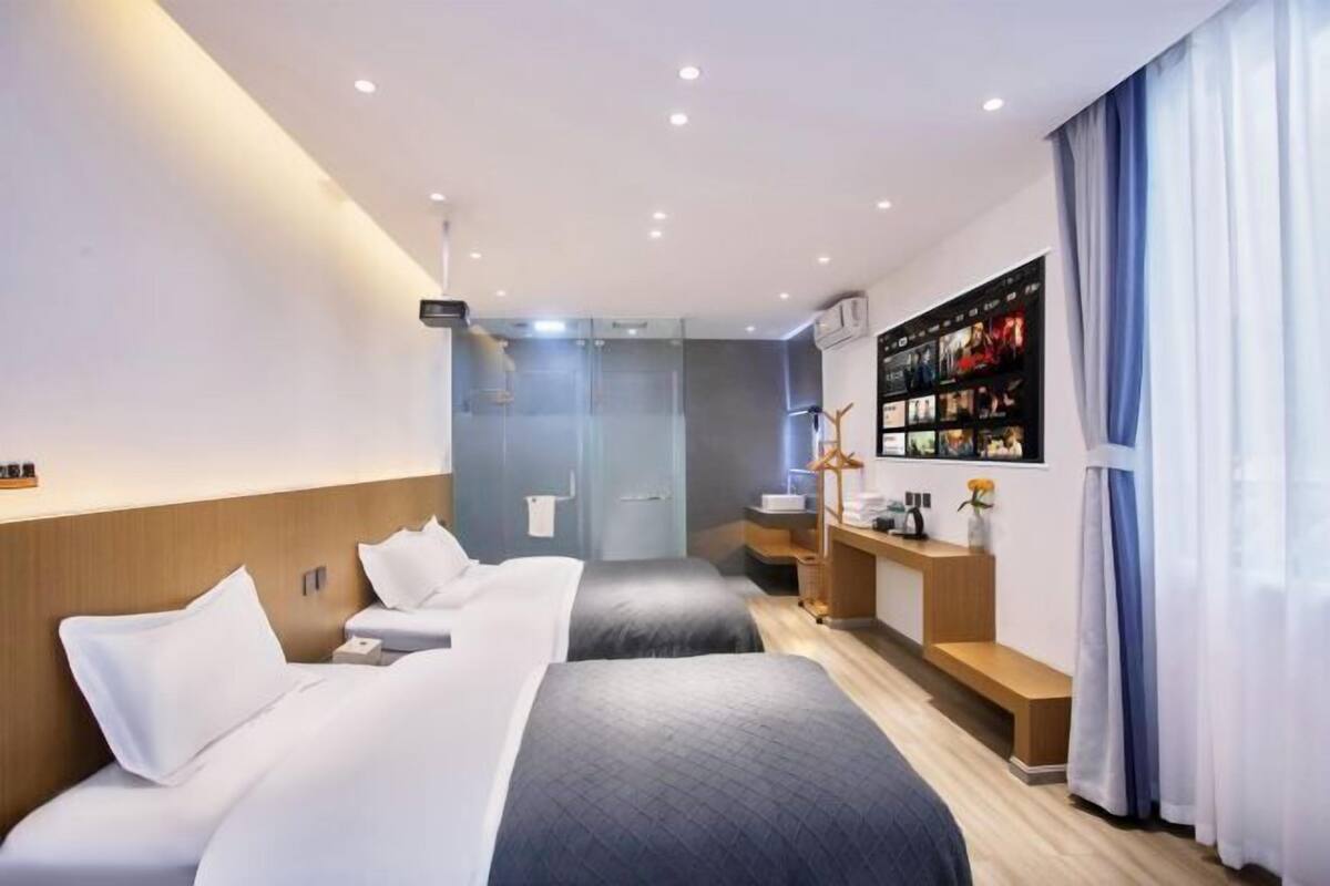 Quarto de hotel com duas camas, uma toucador com espelho, um prateleira com artigos de higiene pessoal e uma TV fixada na parede.