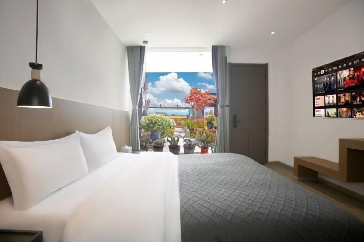 Quarto de hotel moderno com uma cama grande, luminária pendente, vista para uma varanda com plantas e uma televisão fixada na parede.