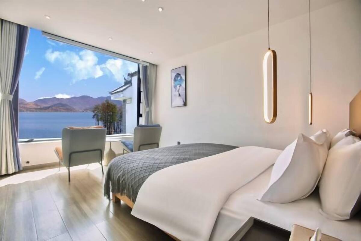 Quarto de hotel moderno com uma cama grande, uma escrivaninha e vista para um lago e montanhas.