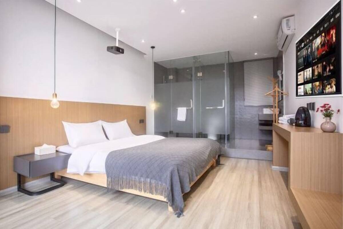 Quarto moderno com cama, mesas de cabeceira, box de vidro e televisão montada na parede.