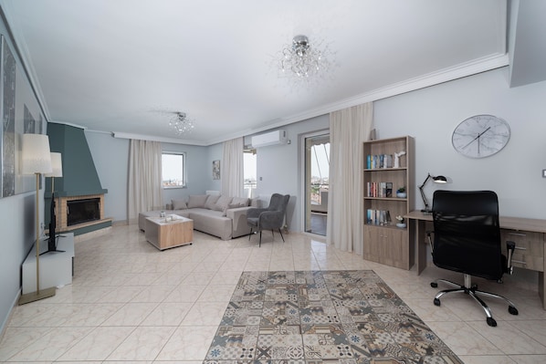 A spacious living room with a fireplace, a sofa, a coffee table, a bookshelf, a desk, and a modern chandelier. - Terminal Square Suites (Markopoulo Mesogaias)