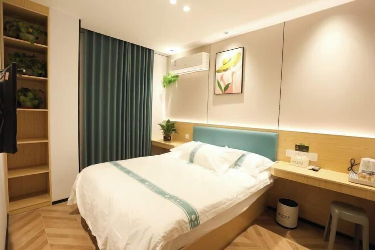 Quarto de hotel com cama, escrivaninha, cadeira, lixeira, prateleira com plantas e ar condicionado fixado na parede.