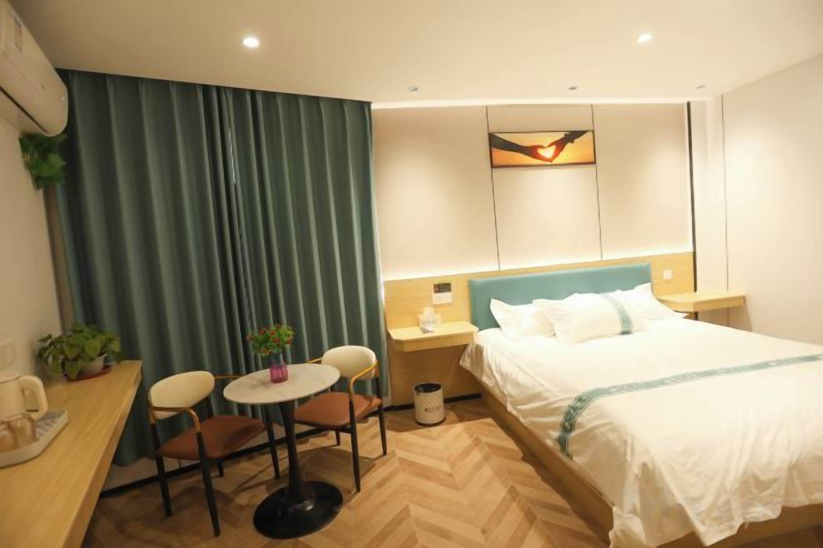 A hotel room with a bed, a desk with a chair, a small table with a plant, and a nightstand with a lamp.