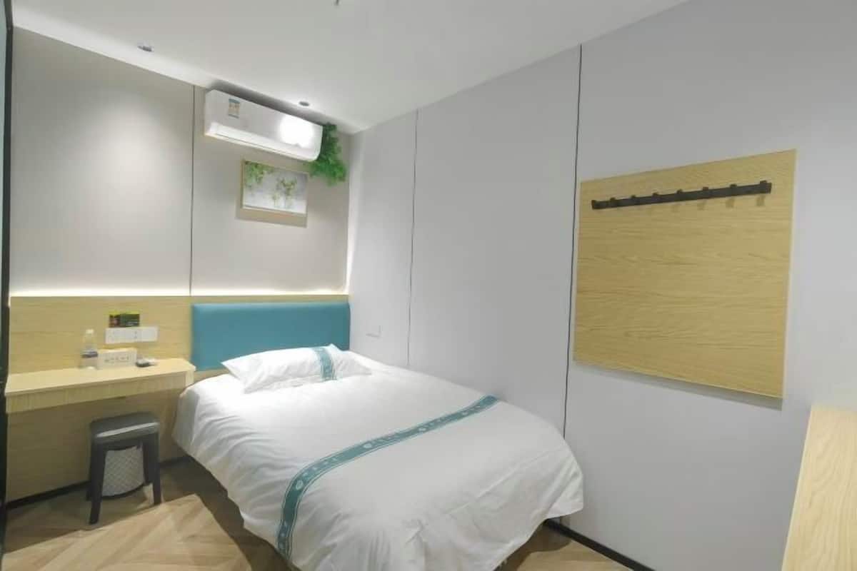 Quarto de hotel com cama, escrivaninha, cadeira, um banquinho pequeno, ar condicionado fixado na parede, quadro emoldurado e painel de madeira na parede.