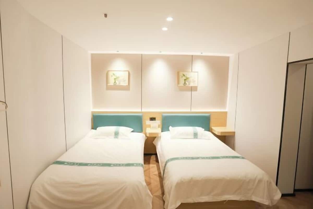 Quarto de hotel com duas camas de solteiro, uma mesa de cabeceira e uma luminária fixa na parede.