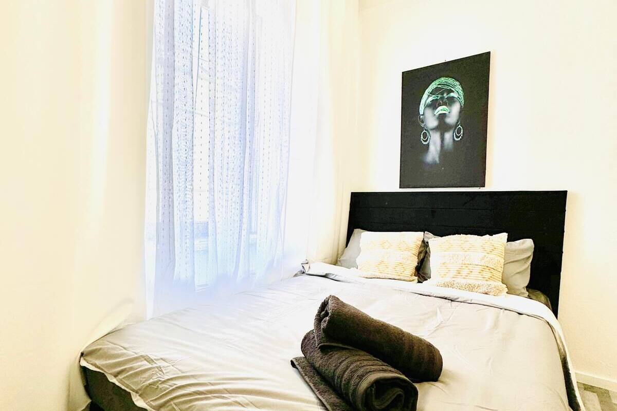 Ein ordentlich bezogenes Bett mit einem zusammengerollten Handtuch, einem schwarzen Kopfteil und einem Gemälde an der Wand.