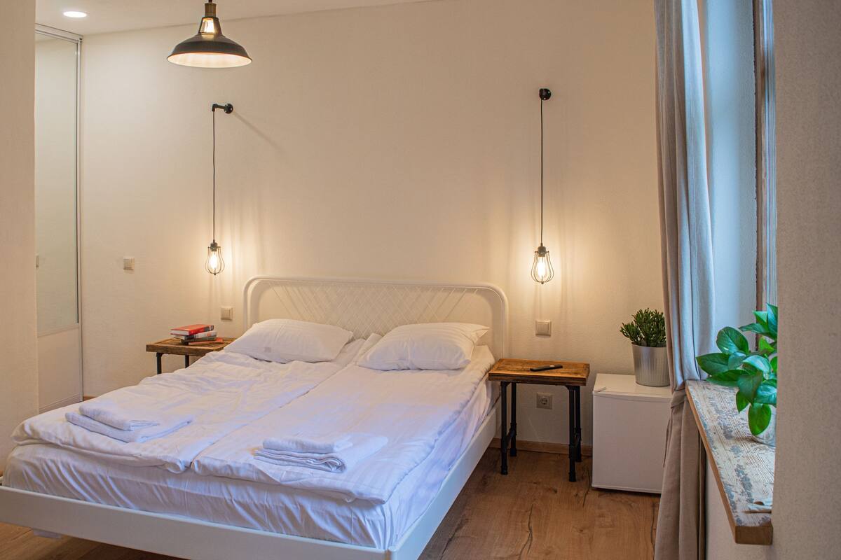 A neatly made bed with white linens, a small bedside table with a book and a potted plant, and a compact white appliance on the floor beside the bed.