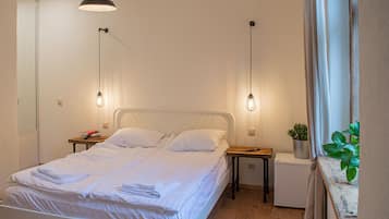 A neatly made bed with white linens, a small bedside table with a book and a potted plant, and a compact white appliance on the floor beside the bed.
