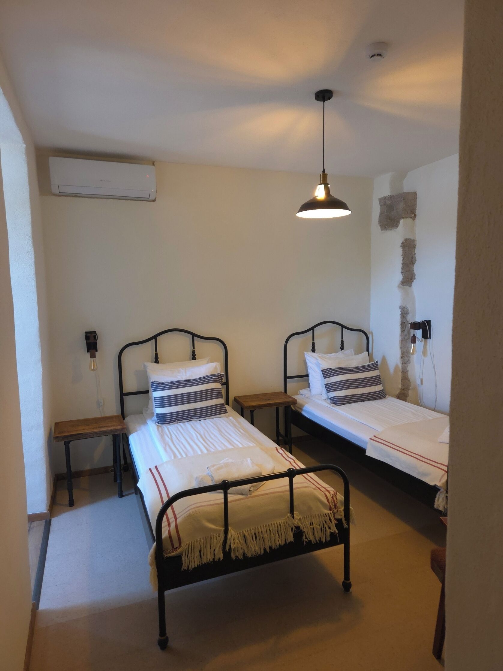 A small, clean room with two single beds, a wooden nightstand, a hanging light fixture, and a wall-mounted air conditioner.