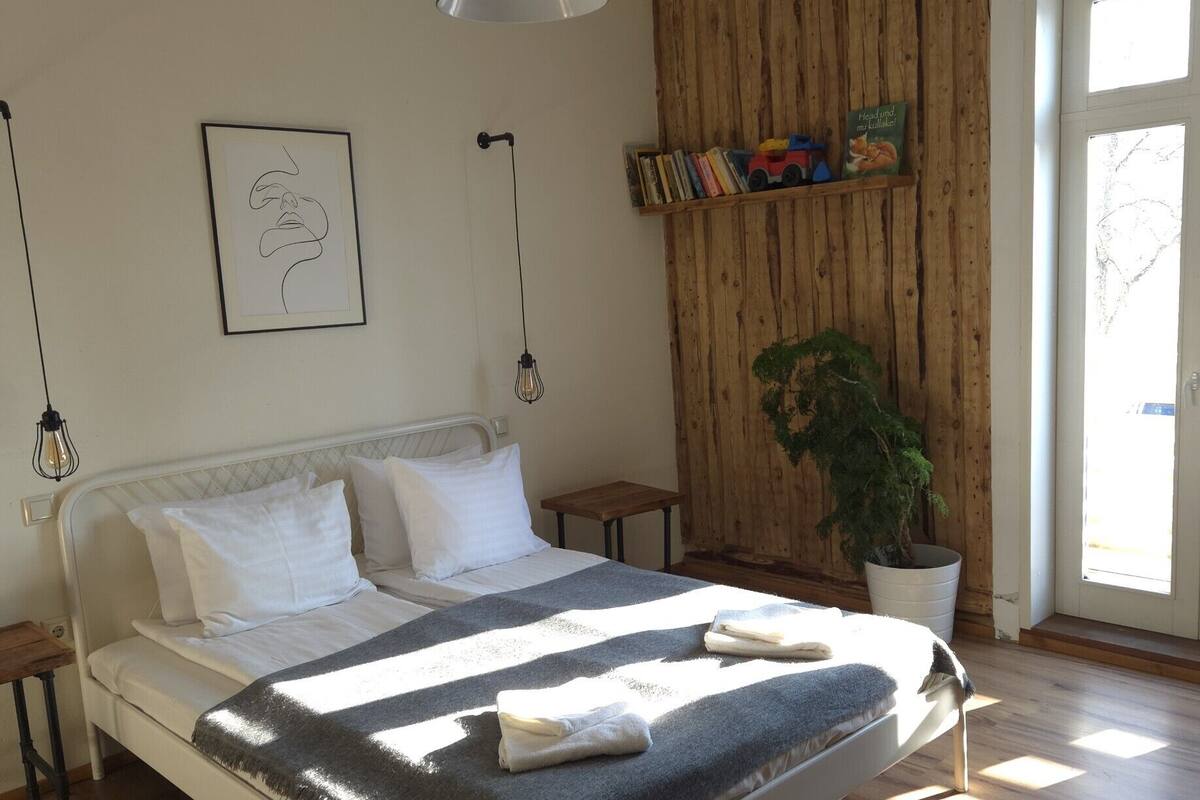 A bedroom with a bed, bedside tables, a hanging light fixture, a framed picture, a wooden wall panel, a bookshelf, and a potted plant.