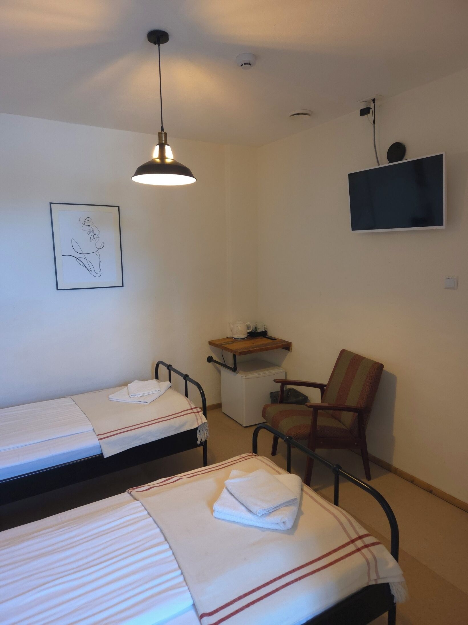 A small, simple room with two single beds, a chair, a TV, and a small shelf with a kettle and cups.