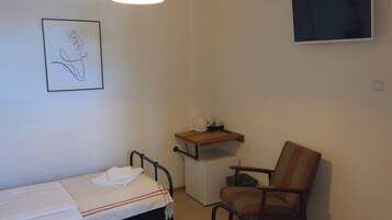 A small, simple room with two single beds, a chair, a TV, and a small shelf with a kettle and cups.