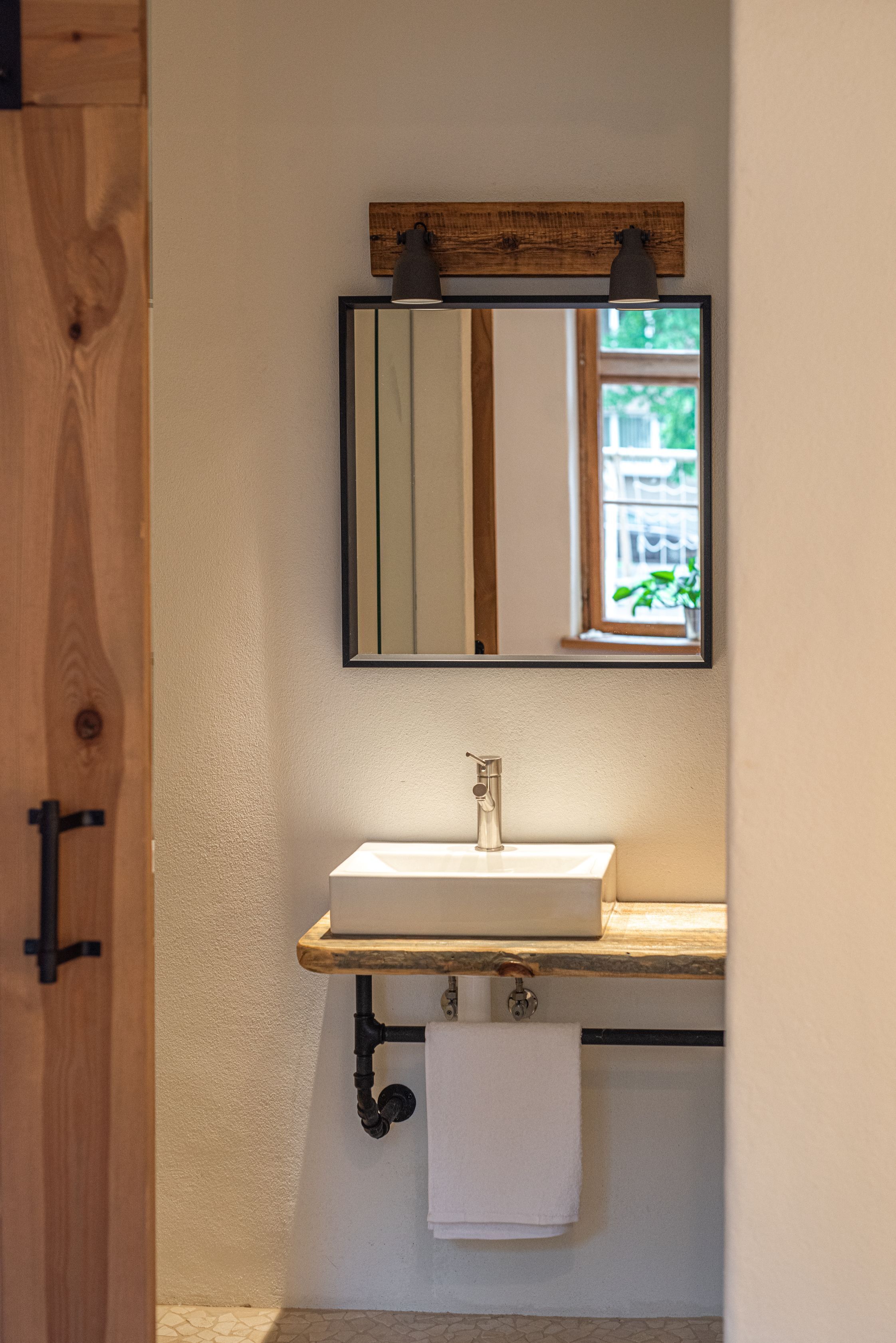 Un baño con puerta de madera, un lavabo blanco, un espejo con marco de madera y una ventana.