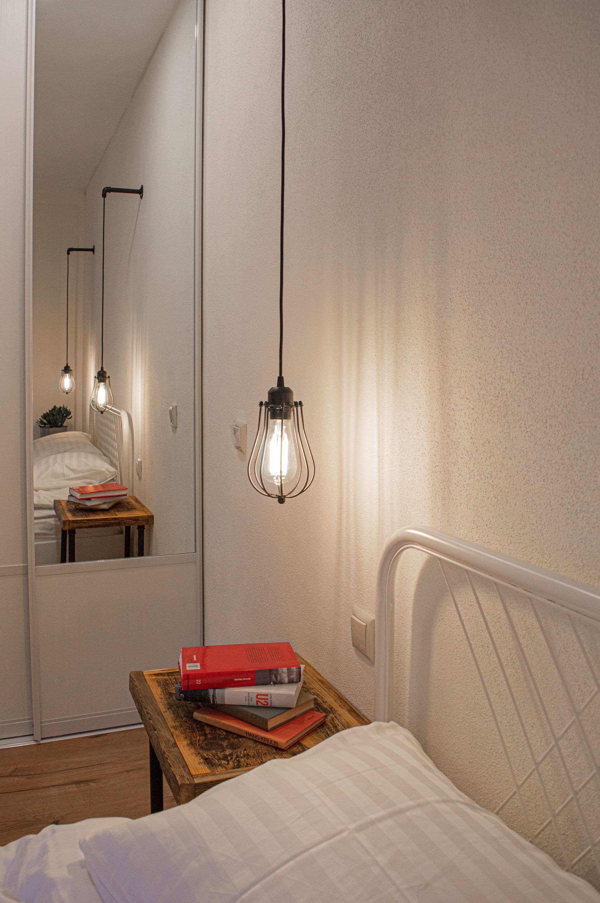 Un dormitorio con cama, mesita de noche con libros, espejo y lámpara colgante.