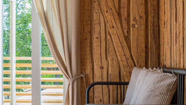A single bed with a metal frame and a striped pillow in a room with wooden walls and a large window with curtains. - TAEAVS  (Võru)