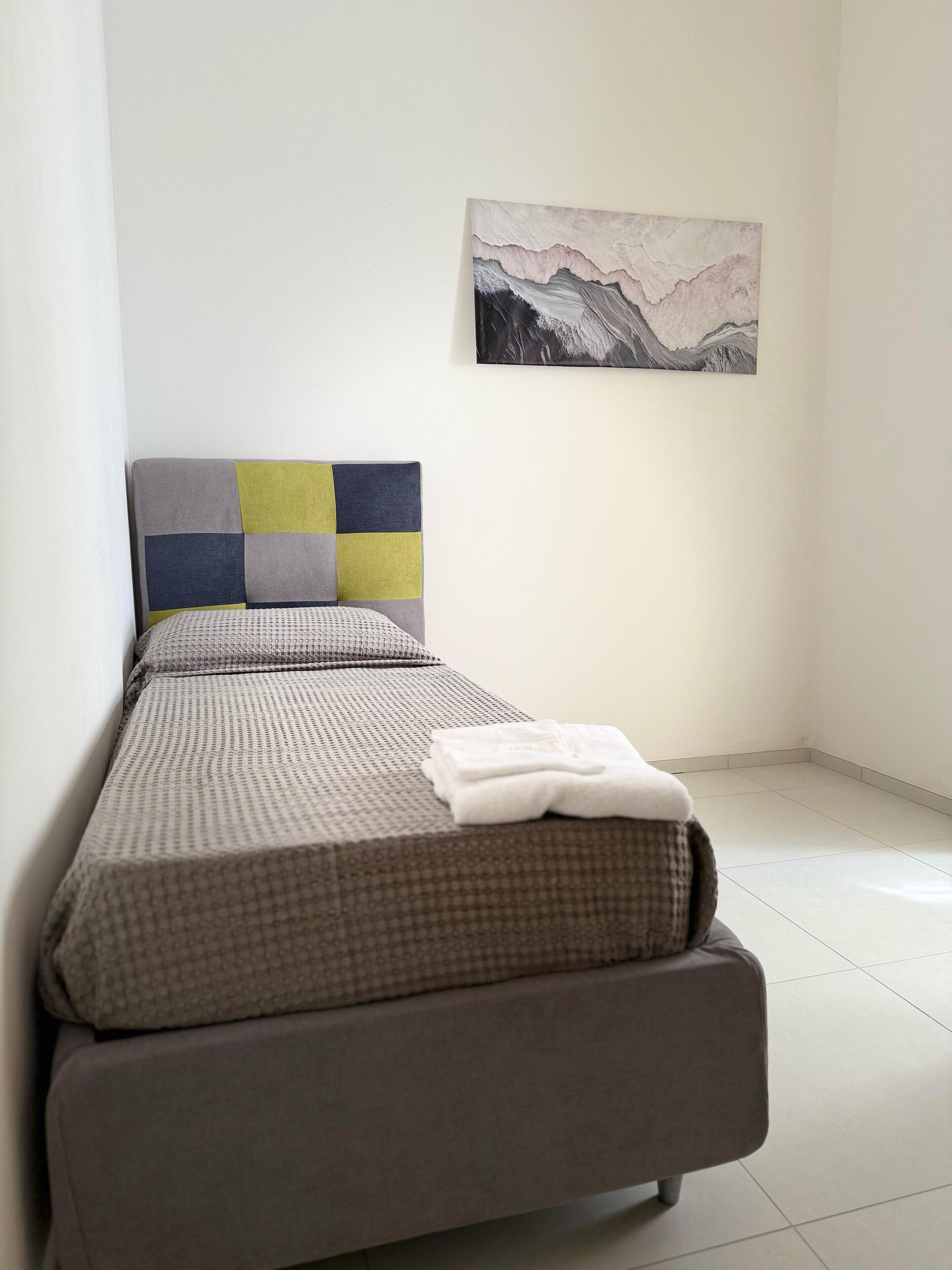Una cama bien hecha con una colcha a cuadros y un cojín con cabecera geométrica. Una toalla blanca doblada está sobre la cama.