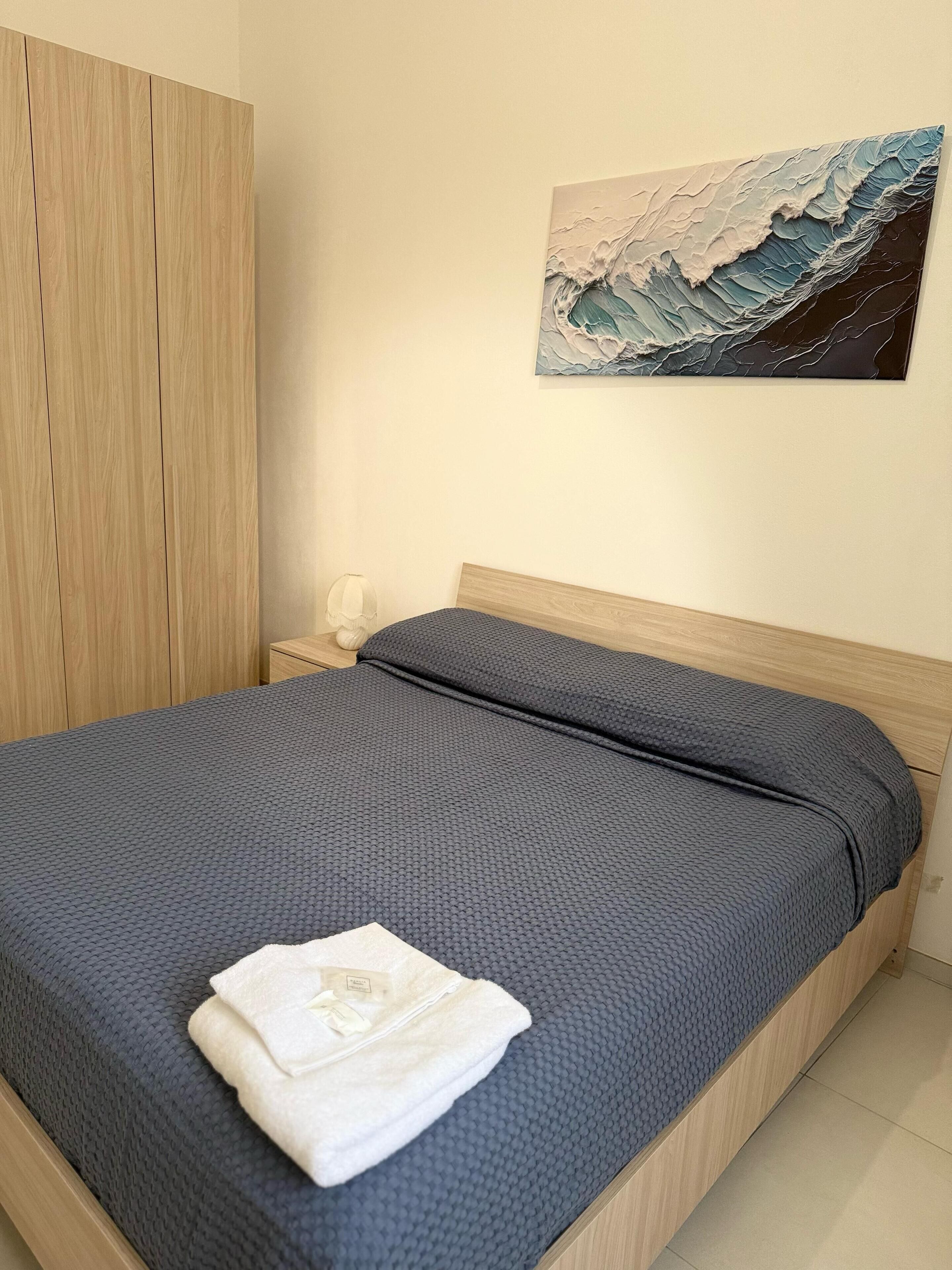 A neatly made bed with a dark textured bedspread and a folded white towel on top.