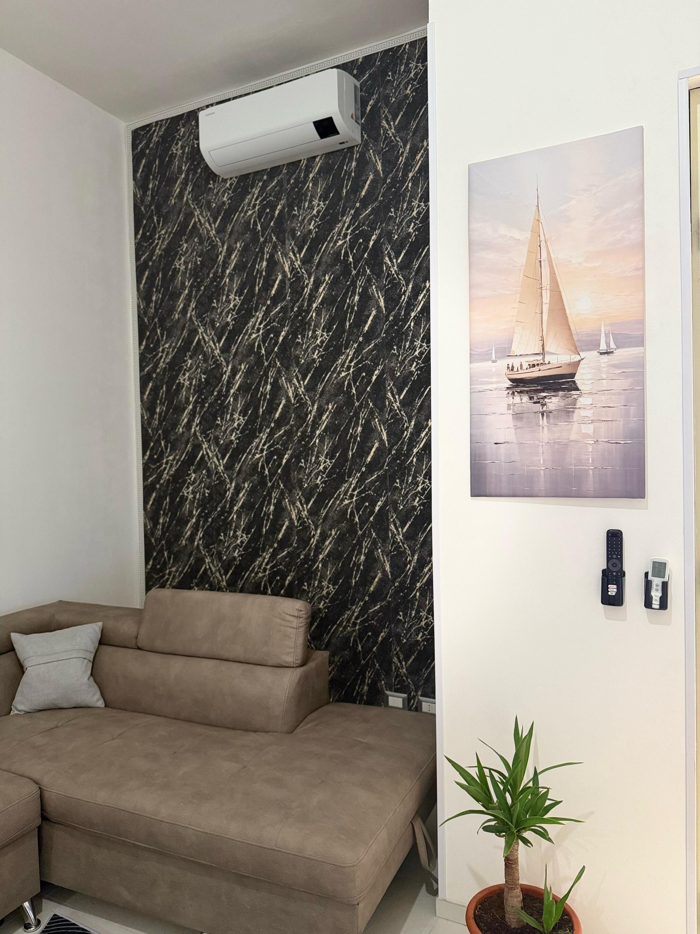 A living room with a sofa, a potted plant, a wall-mounted air conditioner, and a painting of a sailboat.