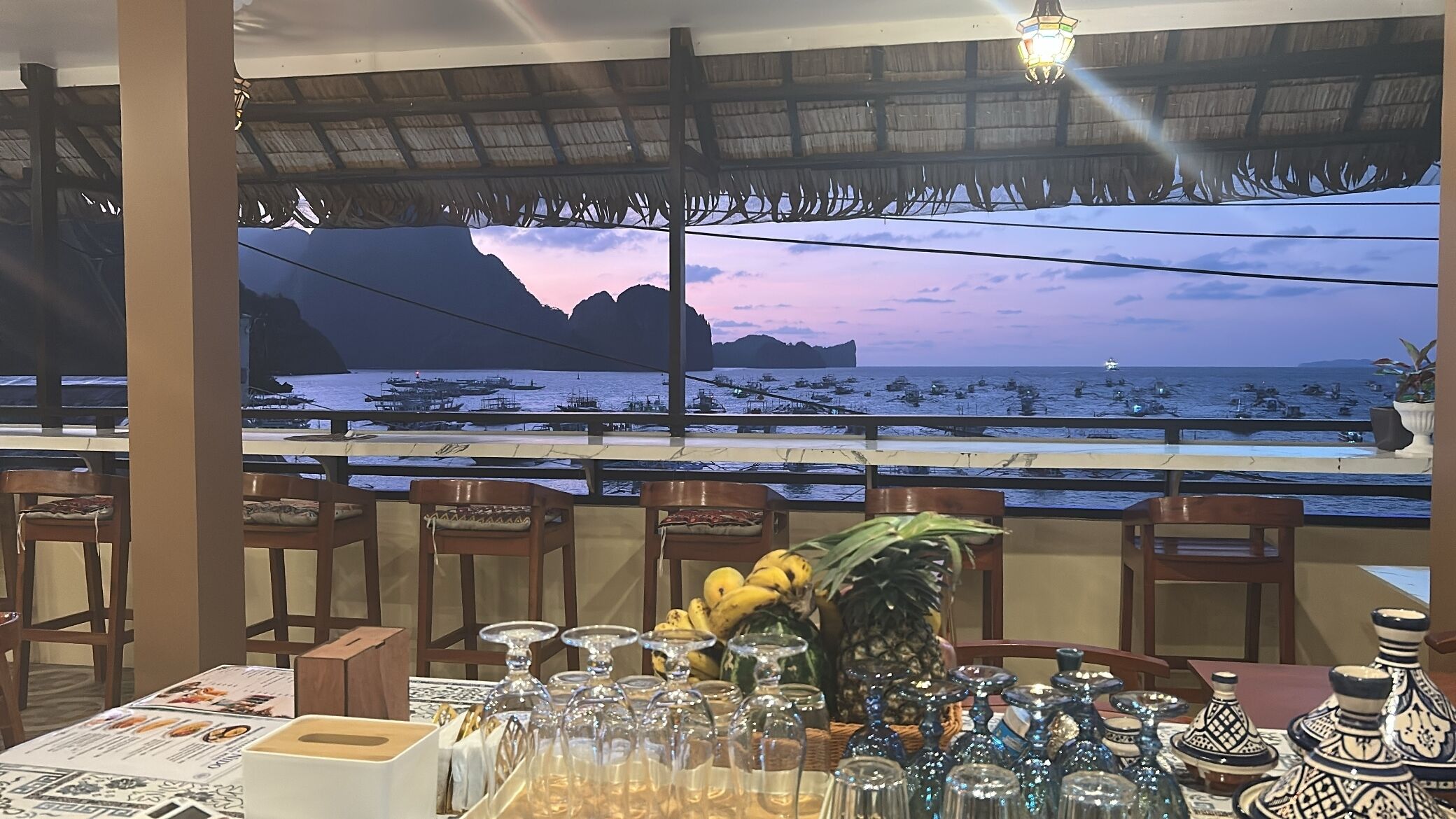 A restaurant with a view of the sea and mountains, featuring wooden chairs, a table with a menu, and decorative items.