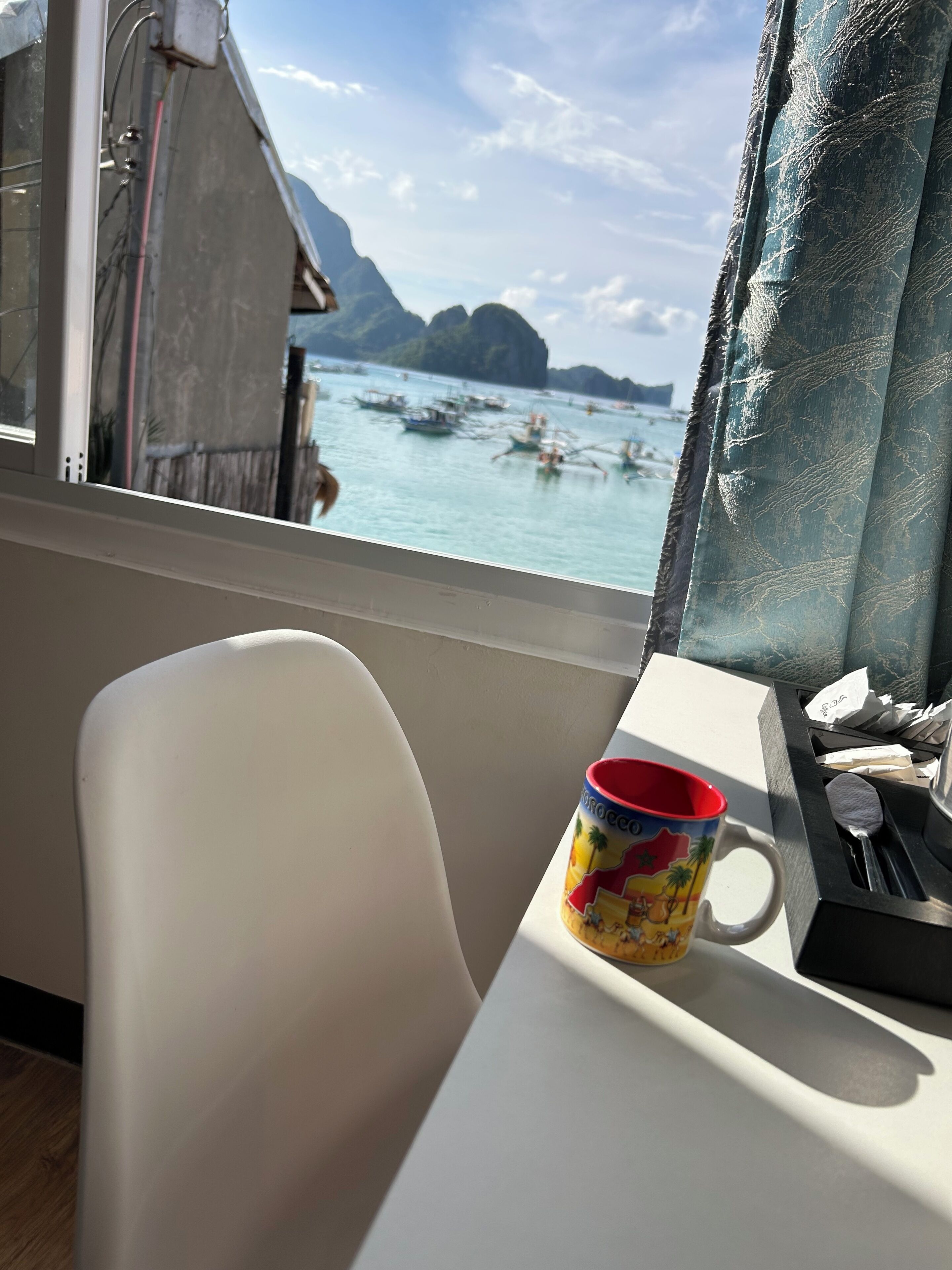 Una habitación con ventana que da a una masa de agua, una silla blanca, una taza de colores y una pequeña mesa con cuchara y soporte para servilletas.