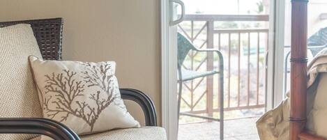 A wicker chair with a cushion, a wooden chair, and a view of an outdoor patio with a railing and chairs.