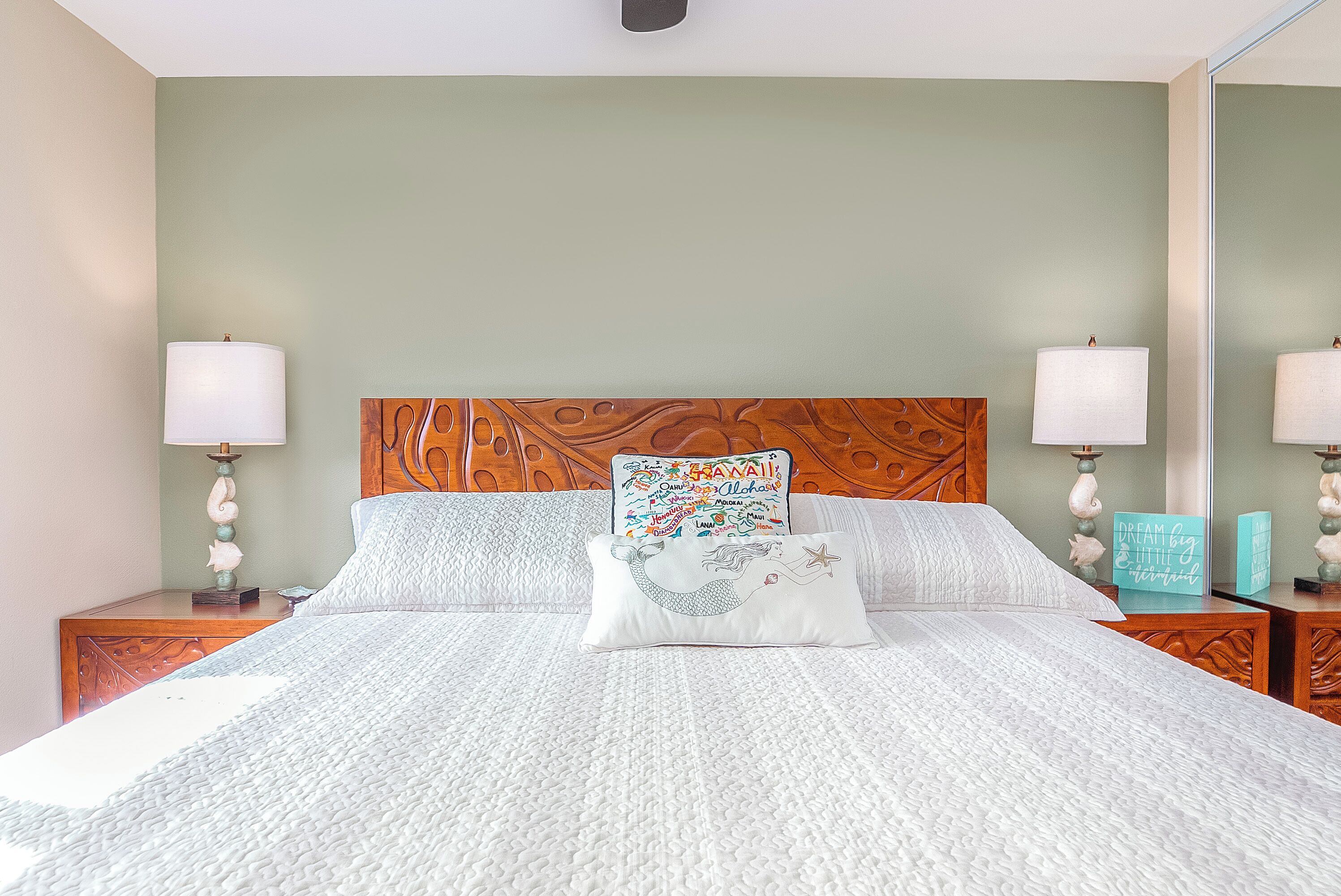 A neatly made bed with a quilted cover, two bedside tables with lamps, a wooden headboard, and a wall with a framed picture.