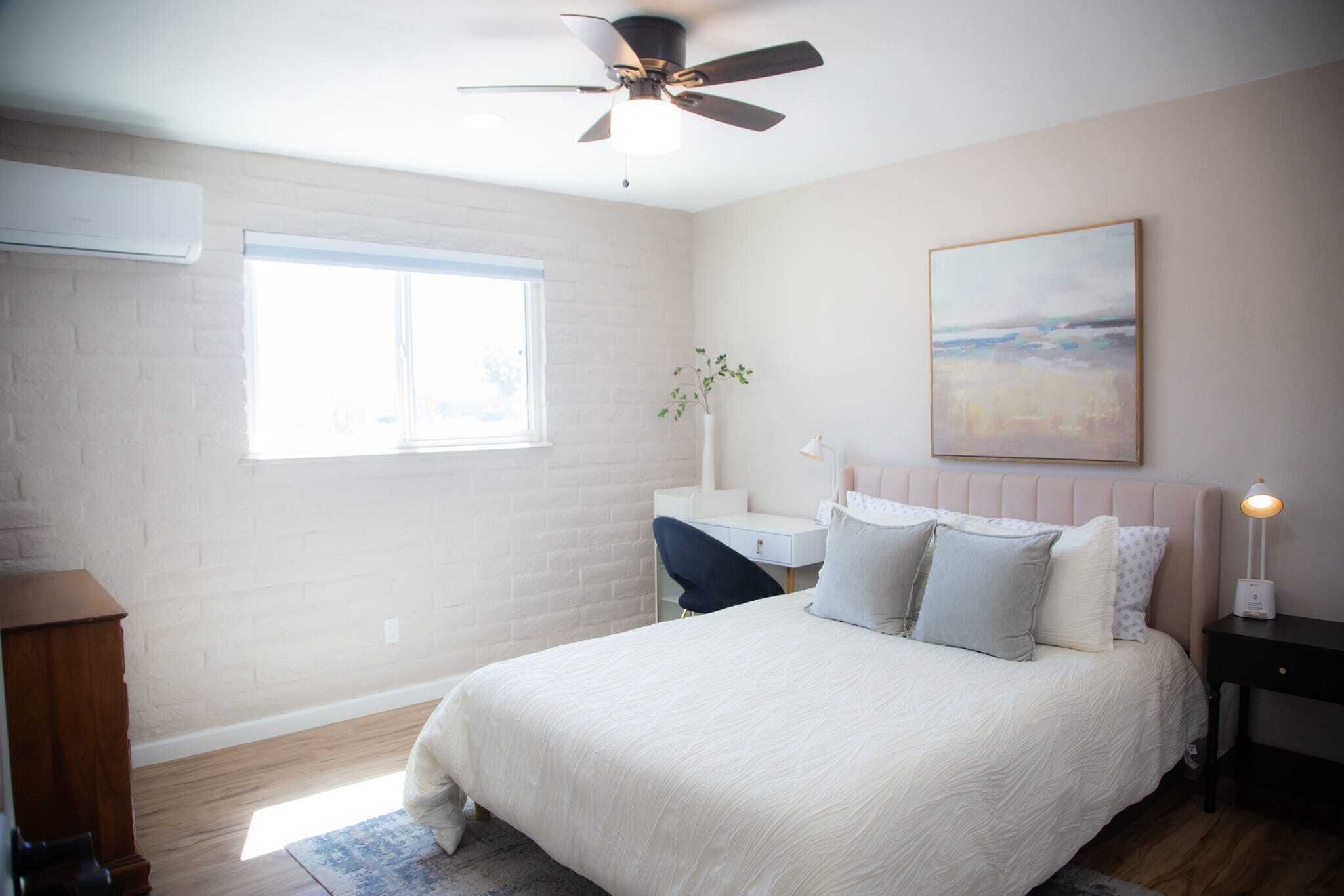 A bedroom with a bed, a desk, a chair, a ceiling fan, a window, and a painting on the wall.