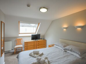 Une chambre à coucher comprenant un lit, une commode en bois, une chaise, un téléviseur posé sur la commode, une fenêtre et deux lampes fixées au mur.