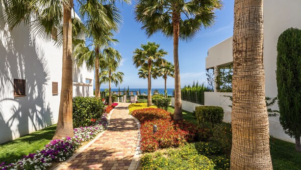 A pathway lined with palm trees and colorful flower beds leading to a building with a view of the sea. - Apartamento La Isla (Mar de Pulpi) (San Juan de los terreros)