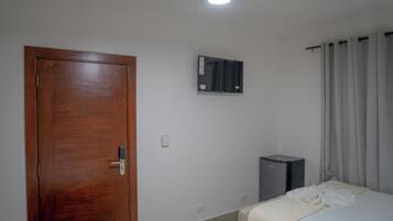 A hotel room with a wooden door, a small desk, a bed with a white bedspread, a mirror on the wall, and a window with curtains.