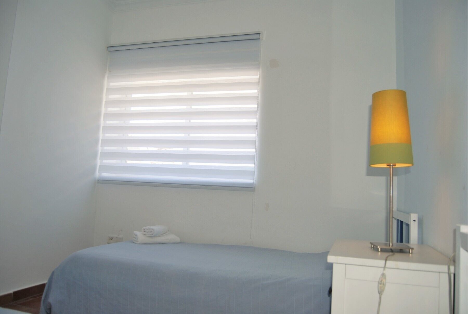 A small, simple room with a bed, a nightstand, a lamp, and a window with blinds.