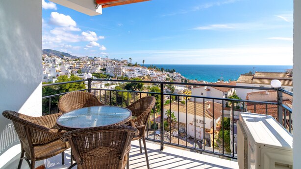 Un balcon doté de meubles en osier et d’une table en verre, offrant une vue sur une ville côtière.