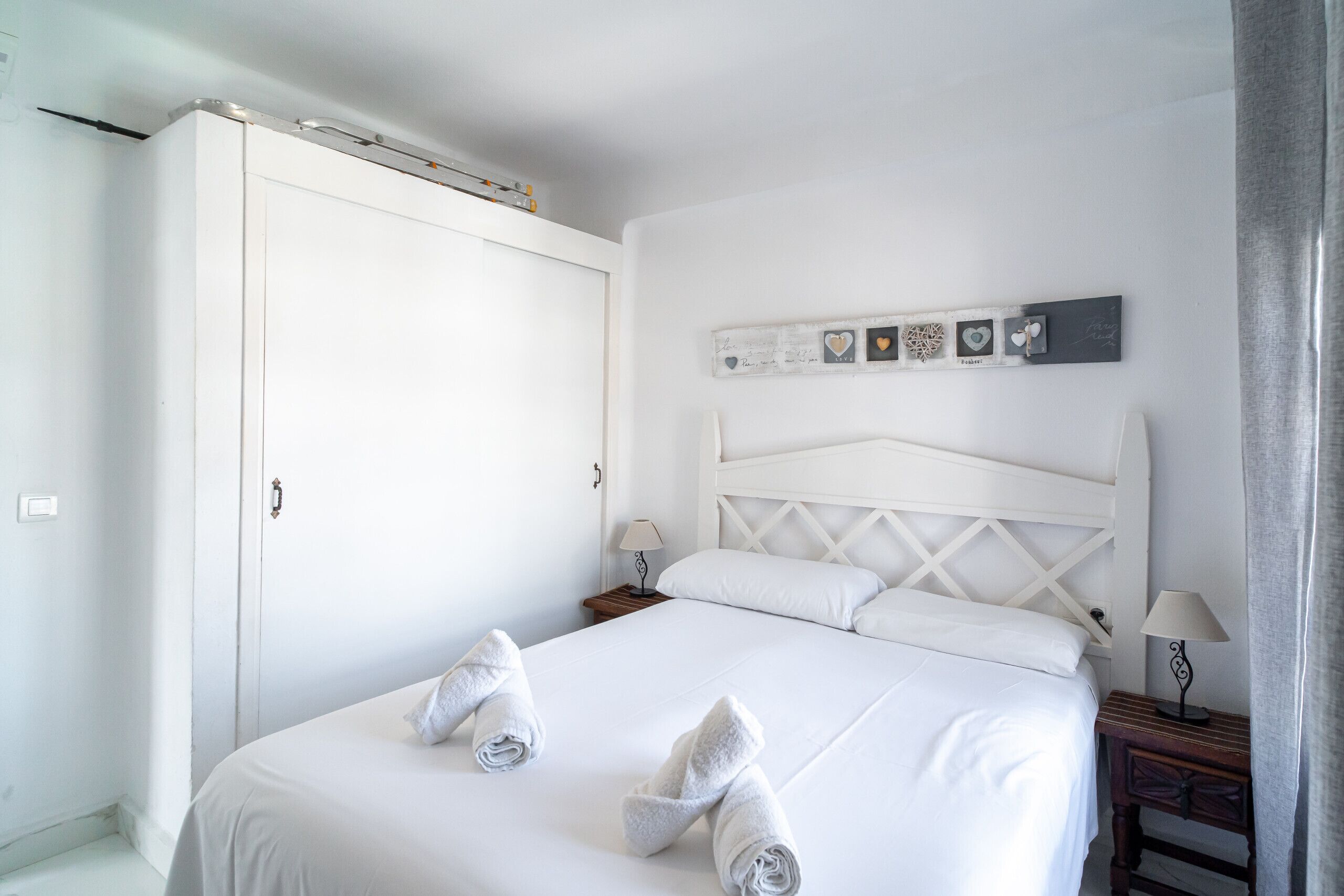 Cama bem arrumada com lençóis brancos e duas toalhas enroladas sobre ela. Um criado-mudo de madeira com abajur fica ao lado da cama. Um guarda-roupa branco é visível à esquerda da cama.