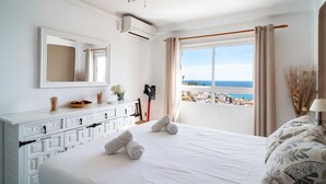 Une chambre d’hôtel avec un lit, une grande fenêtre donnant sur la mer, un ventilateur de plafond et un mini-bar.