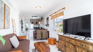 Un salon moderne avec un canapé marron, une table basse en bois et un grand téléviseur. On aperçoit, au fond, une cuisine avec des meubles en bois et un coin repas avec des chaises en osier.