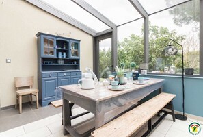 A conservatory with a wooden table set for tea, a blue cabinet with glass doors, and a view of greenery outside.