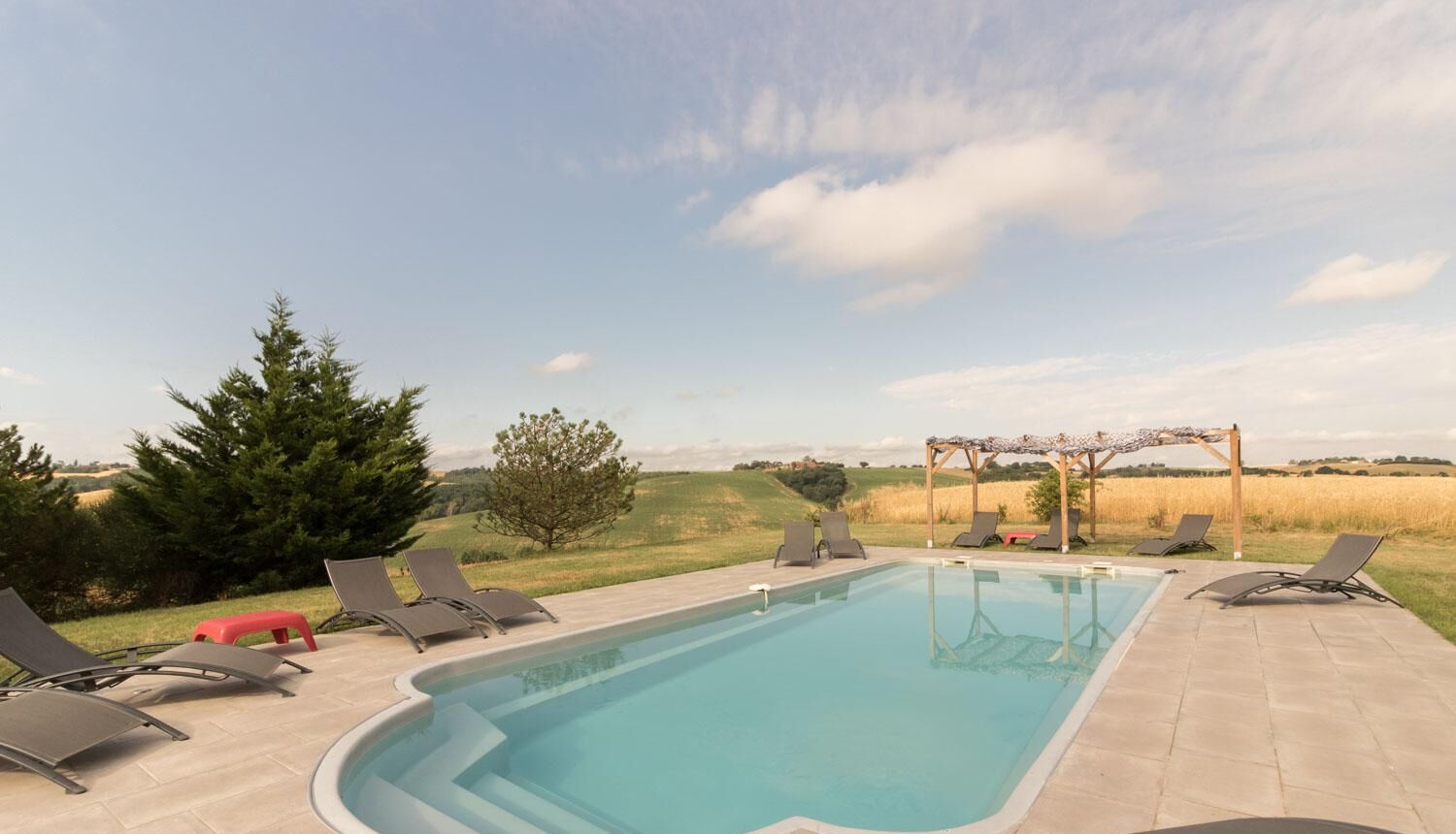 A rectangular swimming pool with a clear blue water surface, surrounded by lounge chairs and a pergola structure.