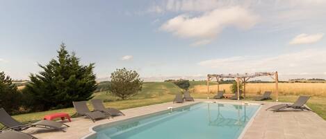 A rectangular swimming pool with a clear blue water surface, surrounded by lounge chairs and a pergola structure.