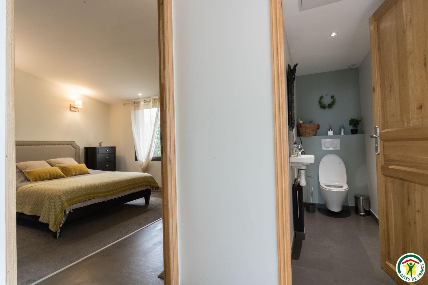 Une chambre à coucher comprenant un lit, une table de chevet et une fenêtre avec des rideaux. La salle de bain est équipée d’un lavabo, de toilettes et d’un espace douche.